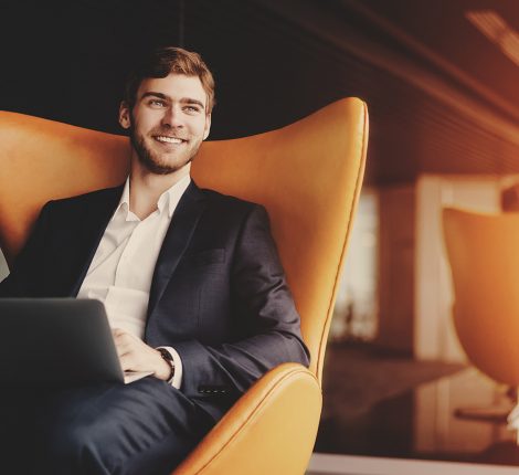 Young smiling successful man entrepreneur in formal business suite with a beard sitting on orange armchair with laptop in luxury office interior